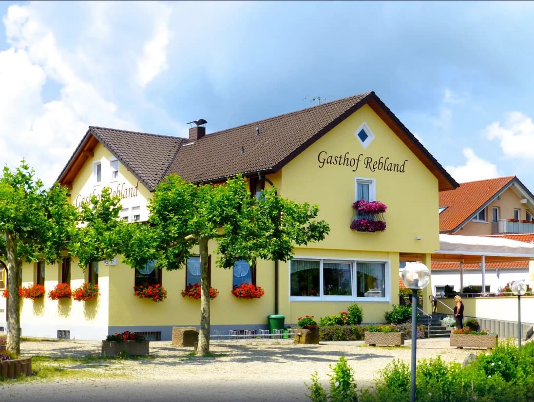 Gelbes Gebäude mit Blumenkästen, grünen Bäumen und einem roten Dach.